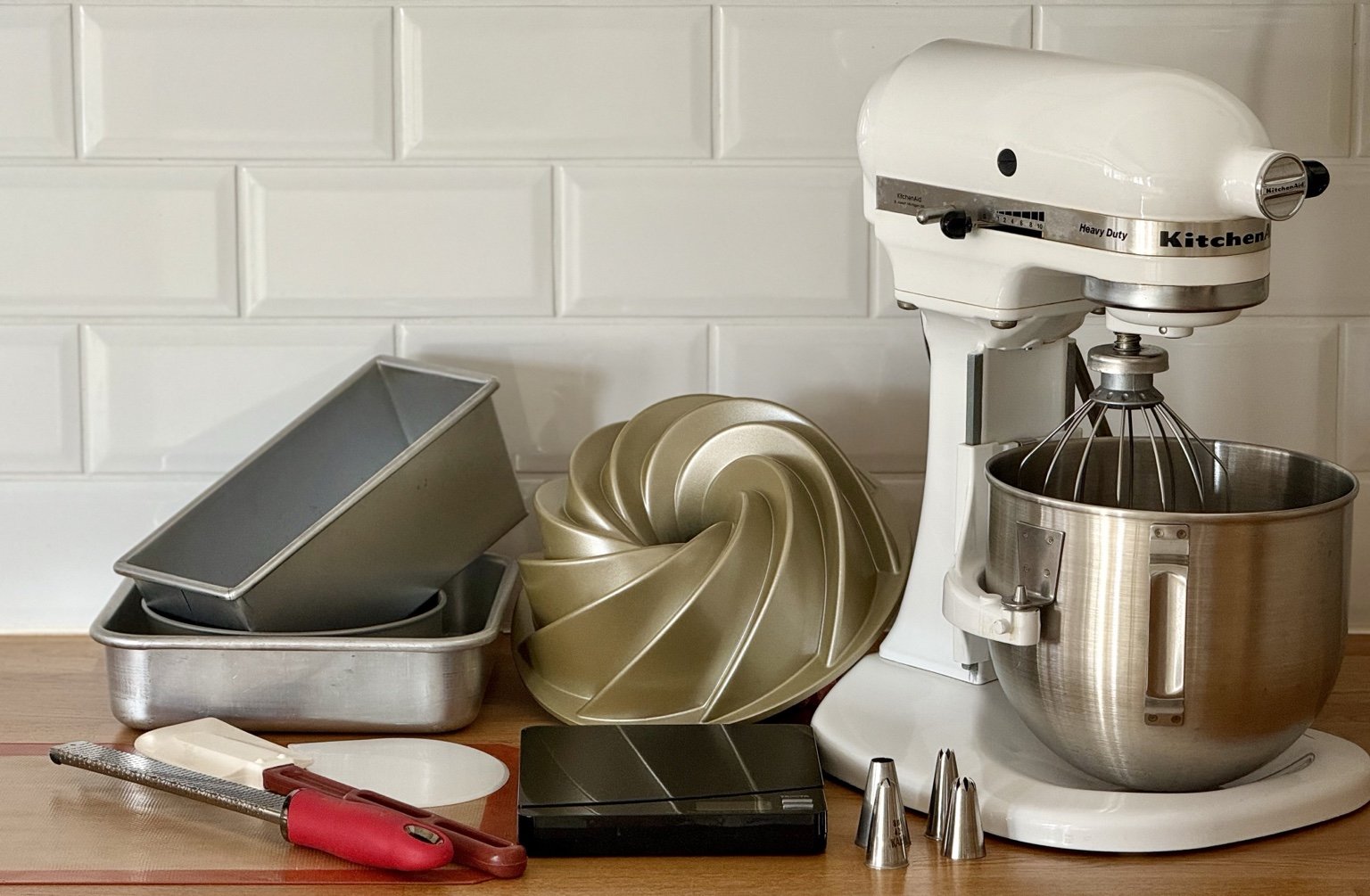 Essential baking tools on wooden kitchen counter including stand mixer, aluminium loaf and cake pans, Bundt pan, digital scale, silicone mat, spatula, bowl scraper and piping tips.