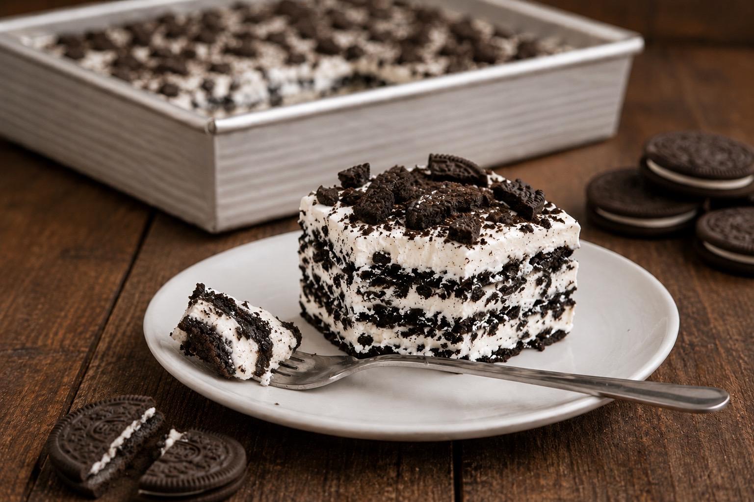 oreo icebox cake slice with layers of whipped cream and chocolate sandwich biscuits on a plate, remaining cake in pan in background